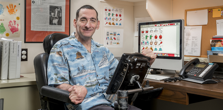 A man with visible cerebral palsy named George Russo sits in an electric scooter equipped with a screen interface. He is wearing a blue Hawaiian shirt, and smiling at the camera.