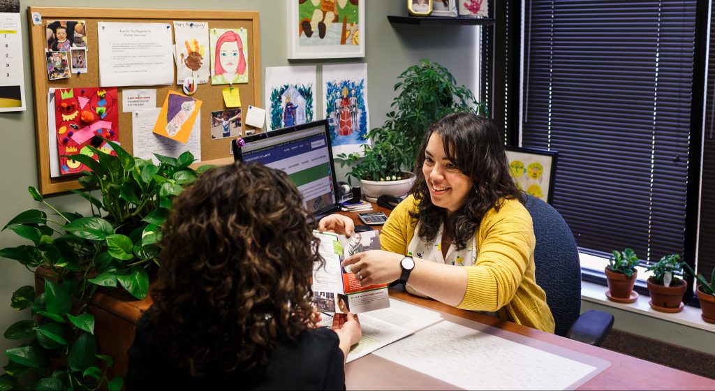 A woman is smiling while leaning over her desk. She is wearing a yellow shawl over her blouse.