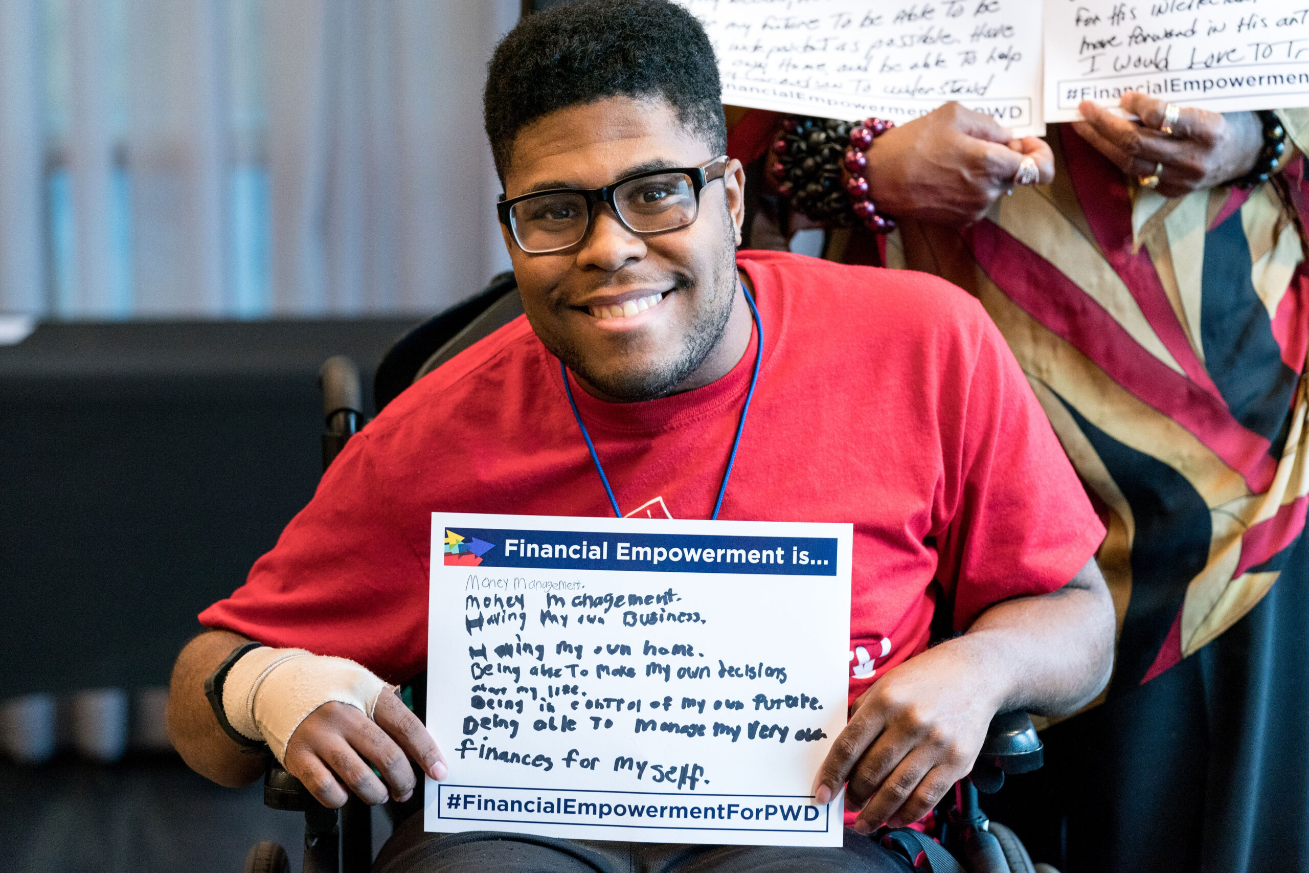 A young man sitting in a wheelchair is smiling and looks at the camera. He is holding a piece of paper titled "financial Empowerment is" and in handwriting it says "Financial Empowerment is...money management. Having my own business. Having my own home. Being able to make my own decisions about my life. Being in control of my own future. Being able to manage my very own finances for myself. #financialempowermentforpwd